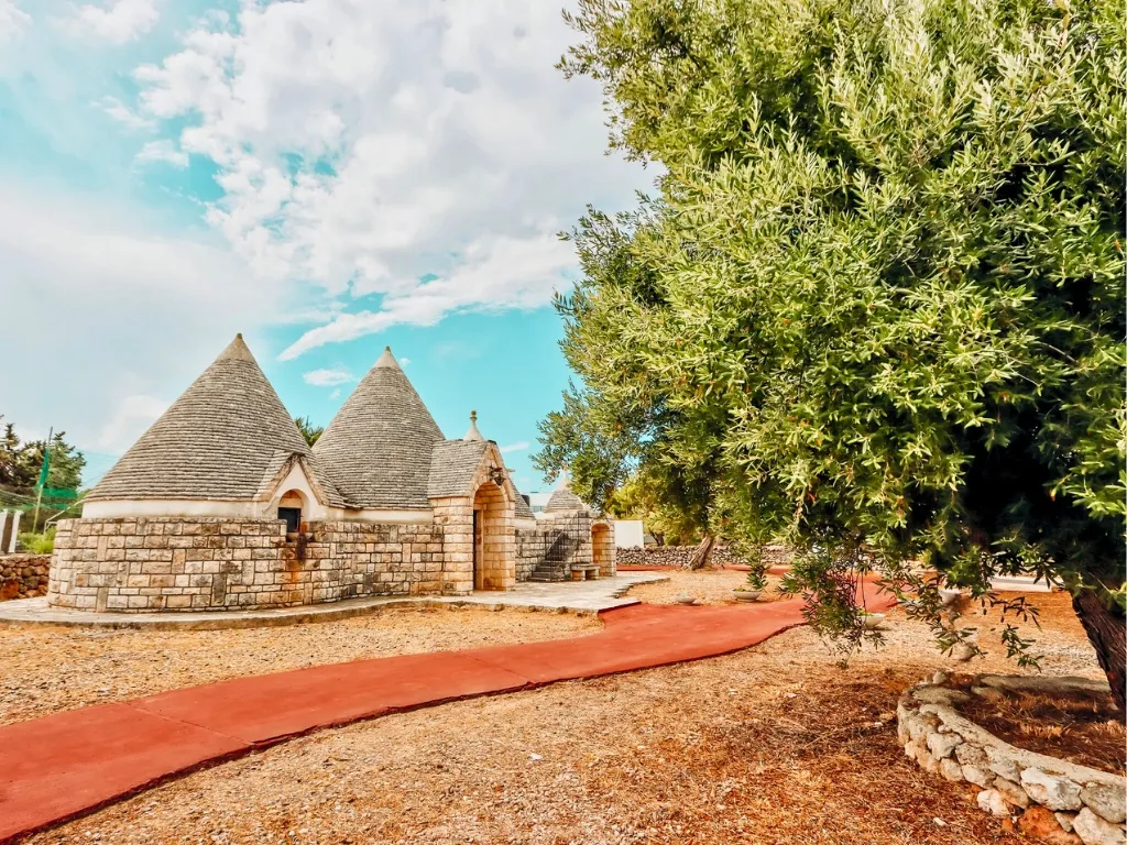 Trulli storici in pietra con vialetto rosso e ulivo secolare