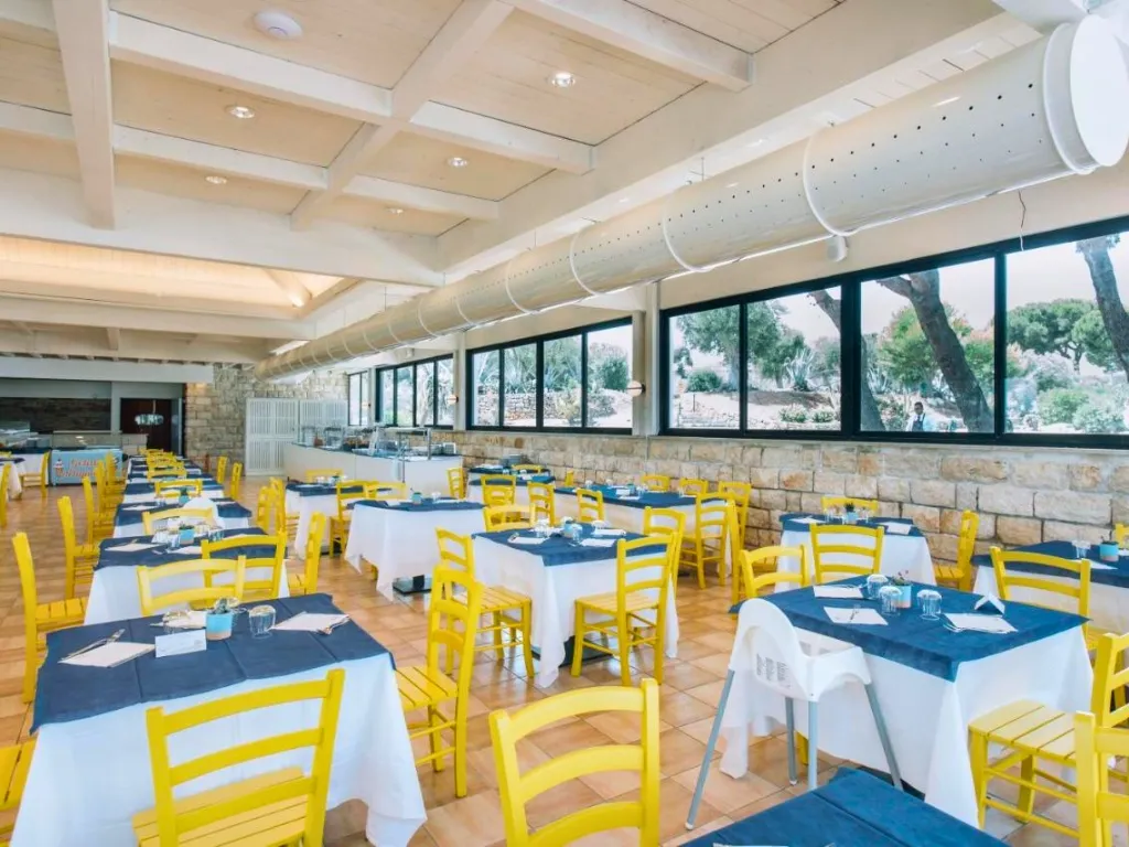 Sala ristorante con sedie gialle, tovaglie blu e parete in pietra con vista sul verde