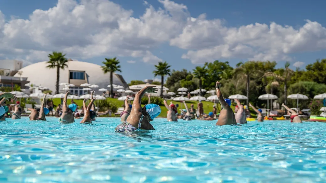 Persone durante lezione di acquagym in piscina con palme sullo sfondo