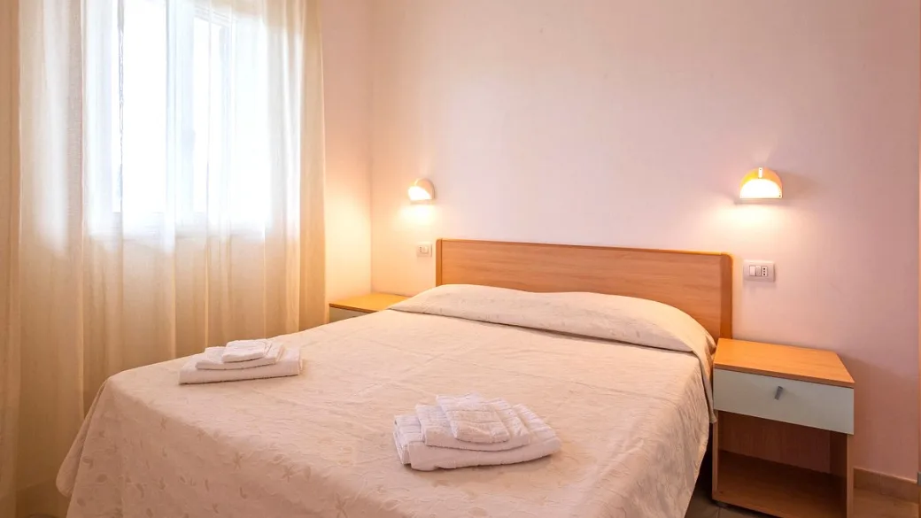 Camera matrimoniale con letto, comodini in legno, applique e finestra con tende leggere