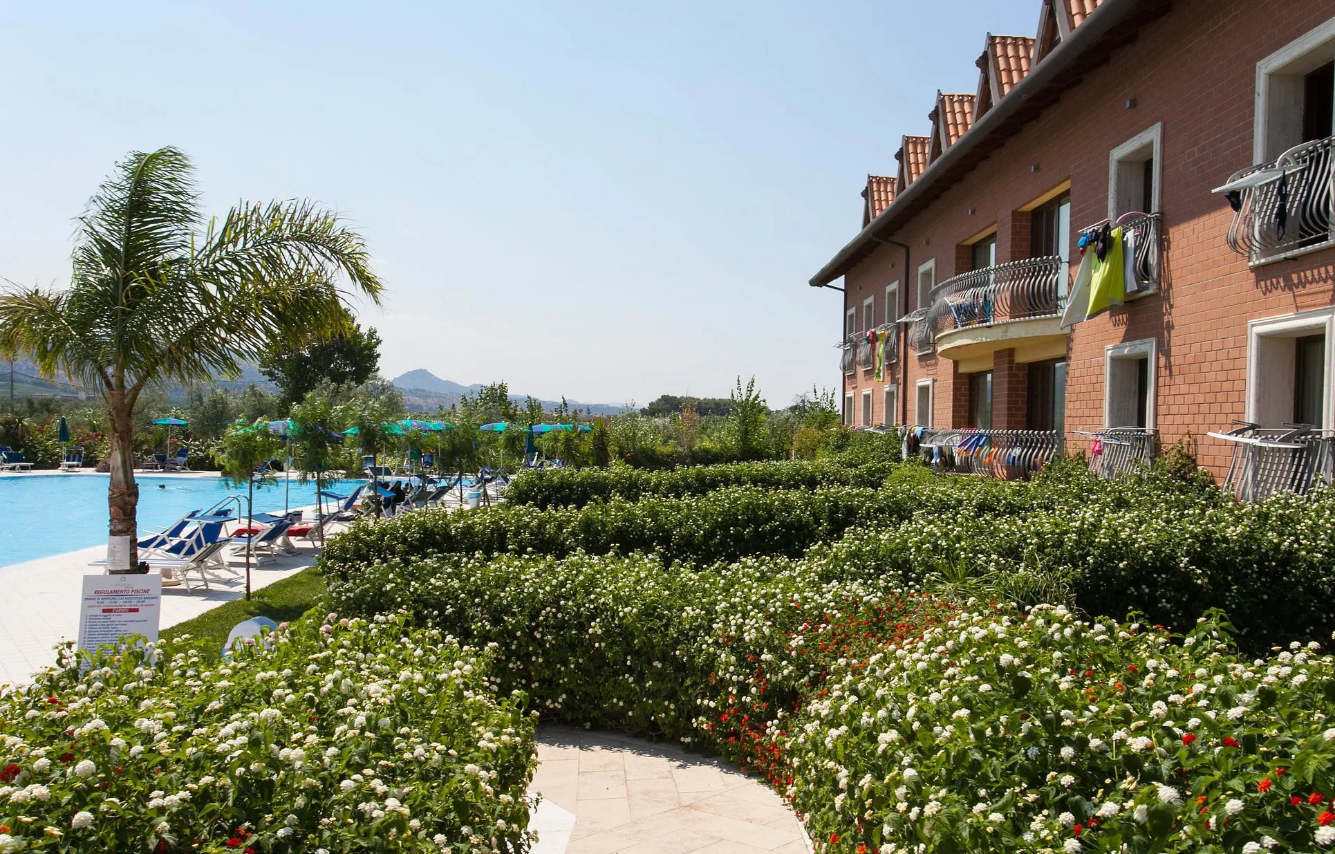 Giardino fiorito con siepi e palme lungo l'edificio del resort e la piscina
