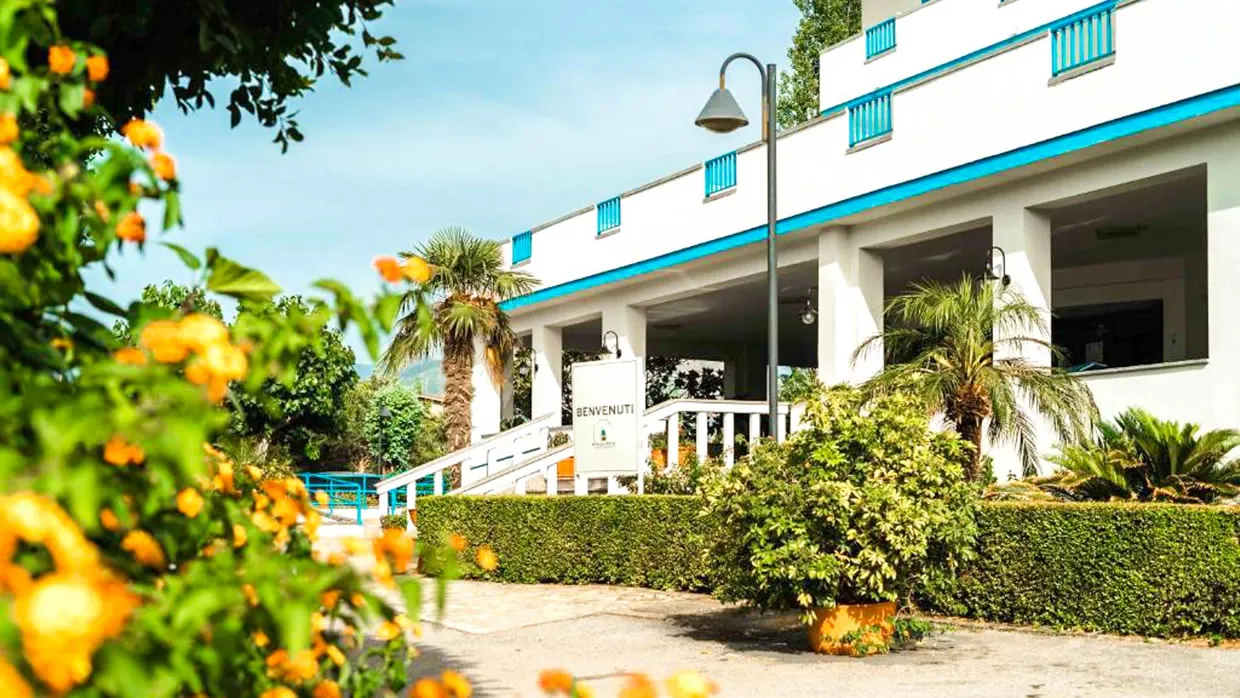 Ingresso dell'hotel con insegna di benvenuto, palme e fiori arancioni