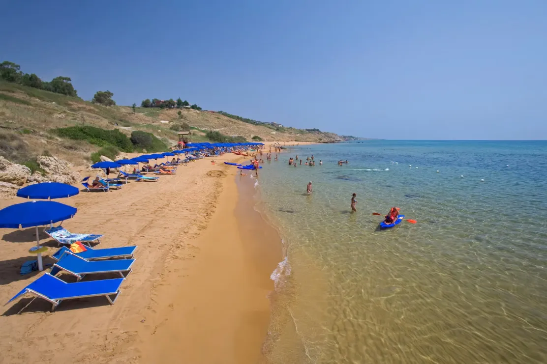 Spiaggia dorata con lettini e ombrelloni blu, bagnanti in acqua e kayak sul mare
