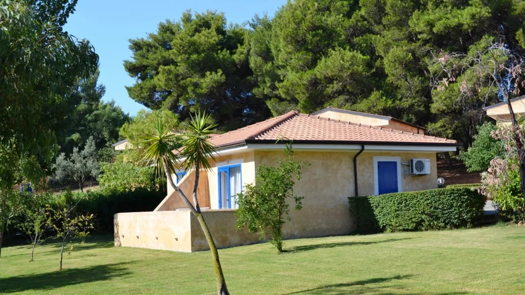 Casetta gialla del resort con persiane blu e giardino curato tra alberi e prato