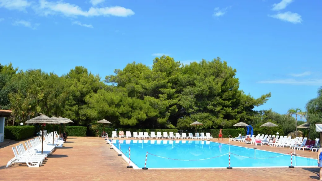 Bordo piscina pavimentato con lettini bianchi, ombrelloni e pineta rigogliosa