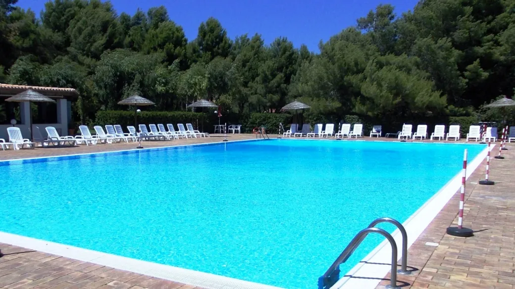 Piscina turchese con lettini bianchi e ombrelloni in paglia circondata da pini