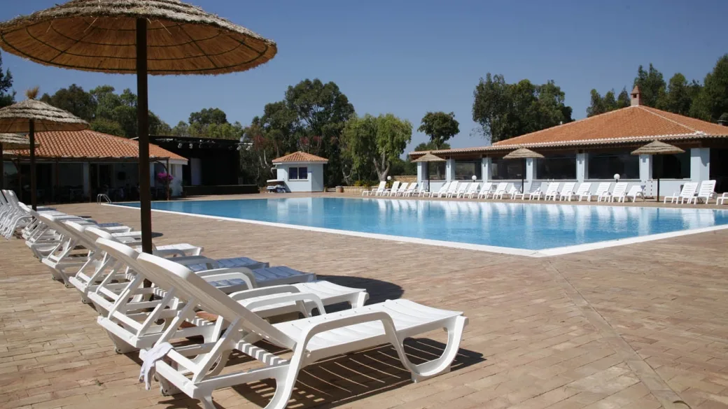 Lettini bianchi e ombrellone in paglia a bordo piscina con edifici del resort sullo sfondo