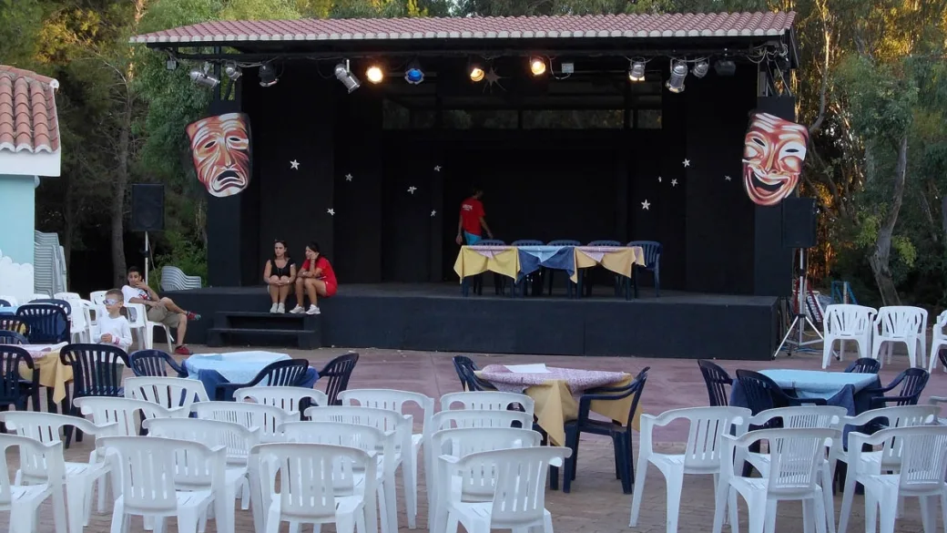 Palco per spettacoli all'aperto con maschere teatrali e platea di sedie bianche