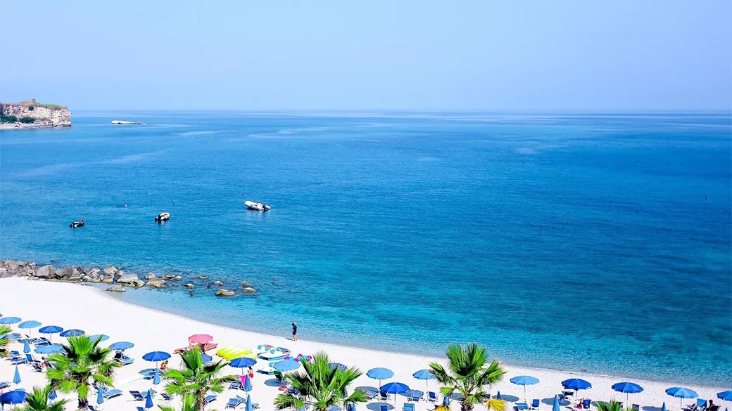 Spiaggia con ombrelloni colorati, palme e mare azzurro con barche all'orizzonte
