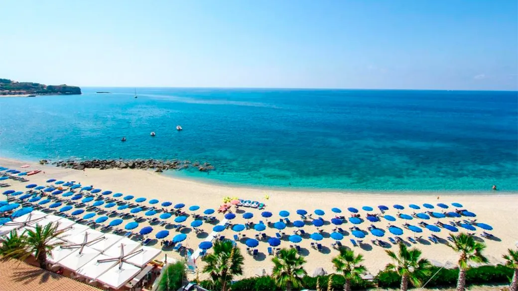 Spiaggia con file di ombrelloni blu, palme e mare cristallino