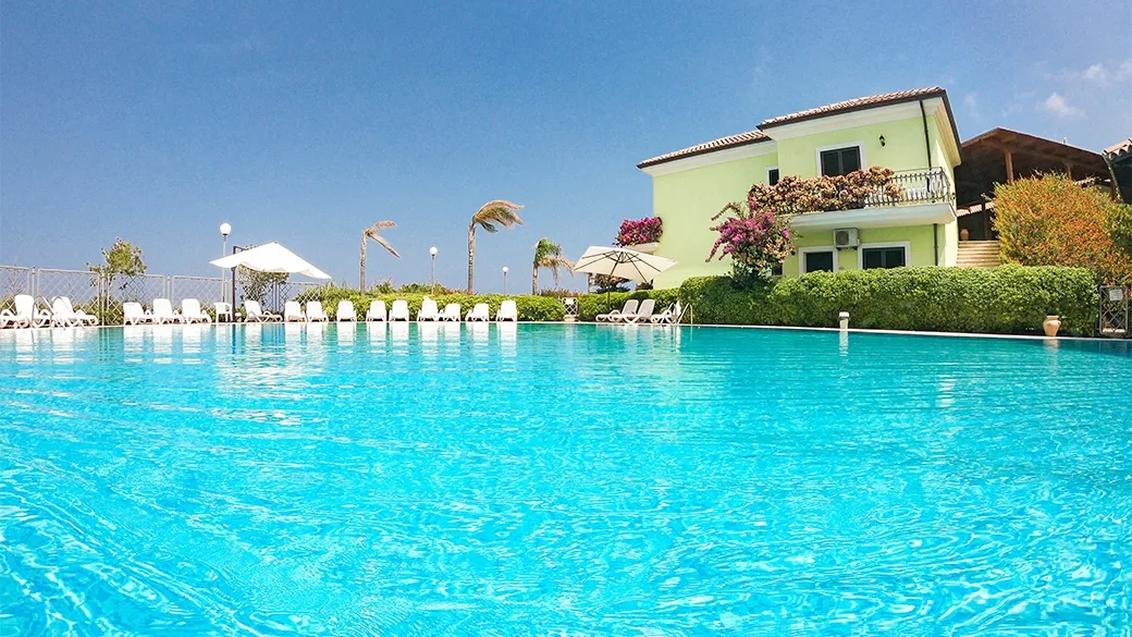 Piscina esterna con lettini e villa colorata sullo sfondo sotto cielo azzurro