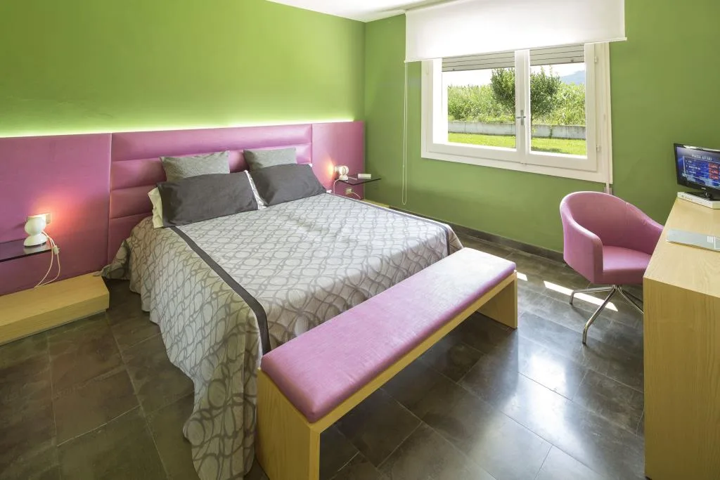 Camera colorata con parete verde, testiera e poltrona rosa, letto matrimoniale e finestra sul verde