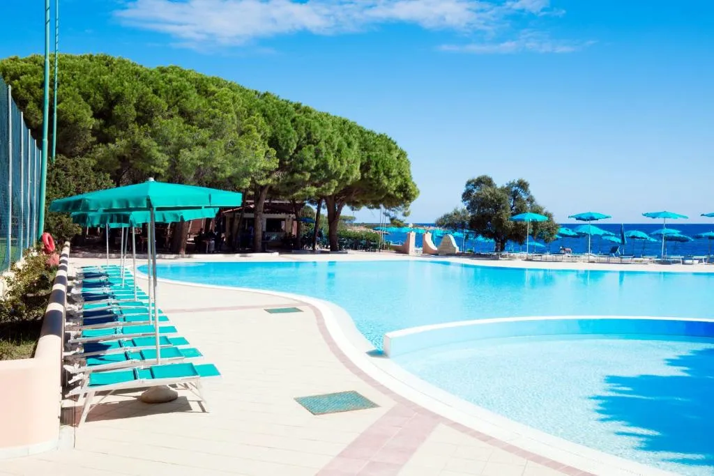 Piscina con lettini blu e ombrelloni verdi tra pini e mare sullo sfondo