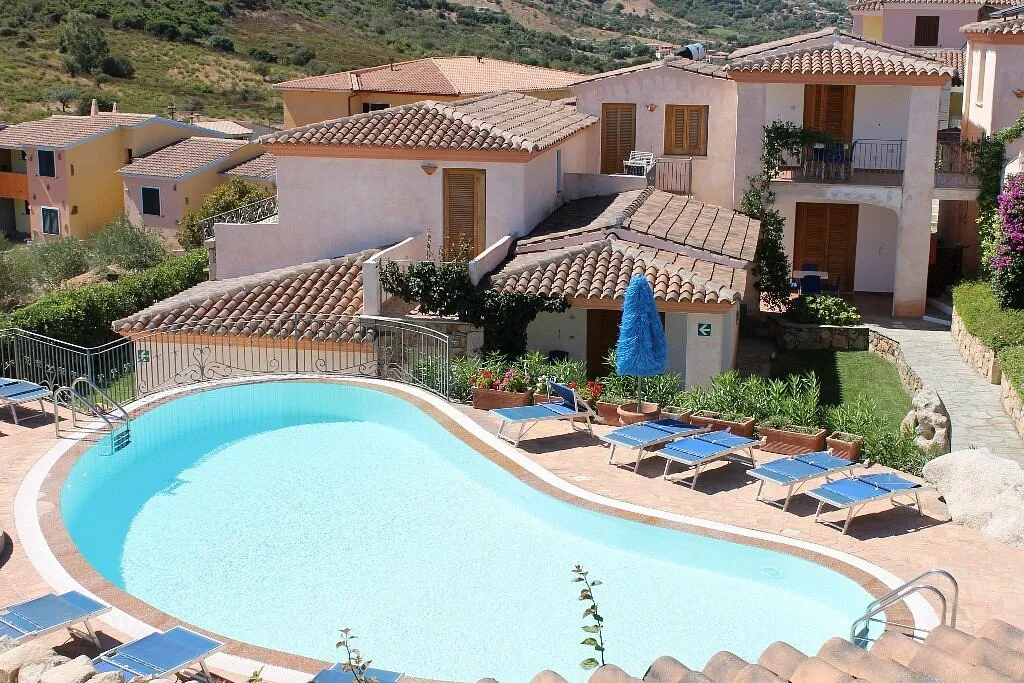Piscina a forma curva con lettini blu e case del residence con tetti in tegole