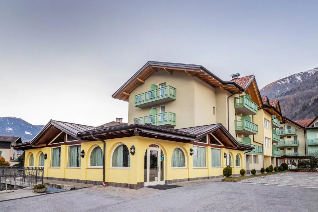 Vista esterna dell'hotel di montagna con facciata gialla, balconi verdi e montagne innevate sullo sfondo