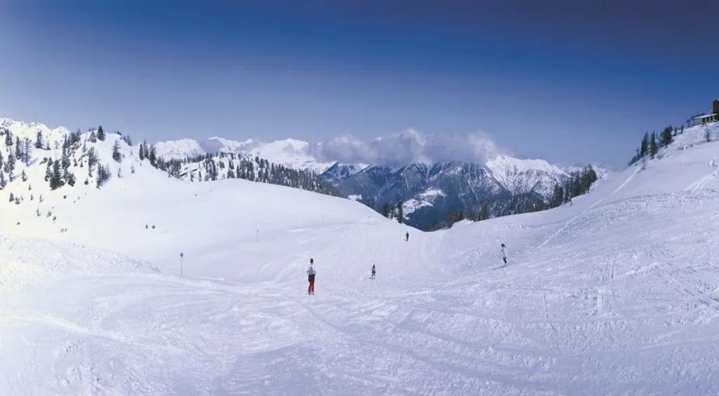 Pista da sci innevata con sciatori e panorama di montagne alpine sotto cielo azzurro