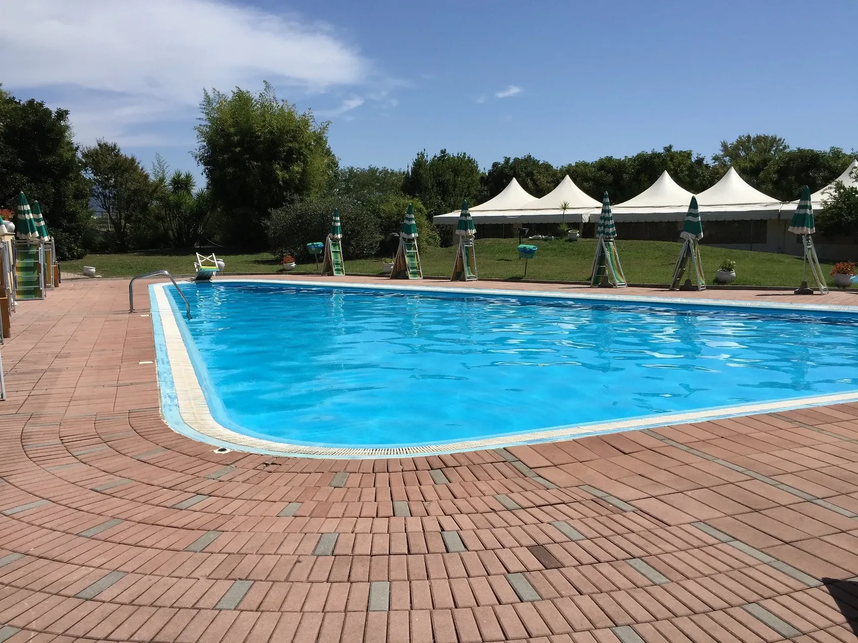 Ampia piscina rettangolare con pavimentazione in cotto, ombrelloni a righe verdi e tendoni bianchi nel giardino