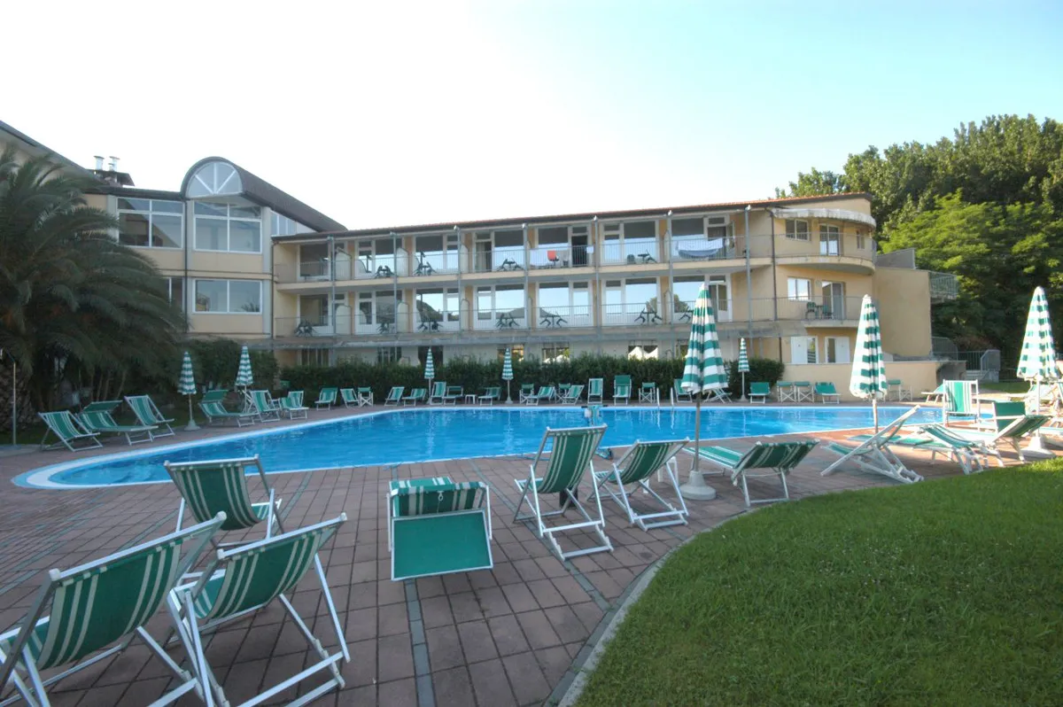 Piscina con numerosi lettini e ombrelloni a righe verdi, edificio dell'hotel a più piani sullo sfondo