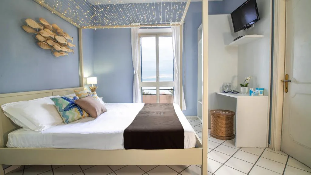 Camera azzurra con letto matrimoniale, luci decorative a soffitto, TV e vista mare dalla porta finestra