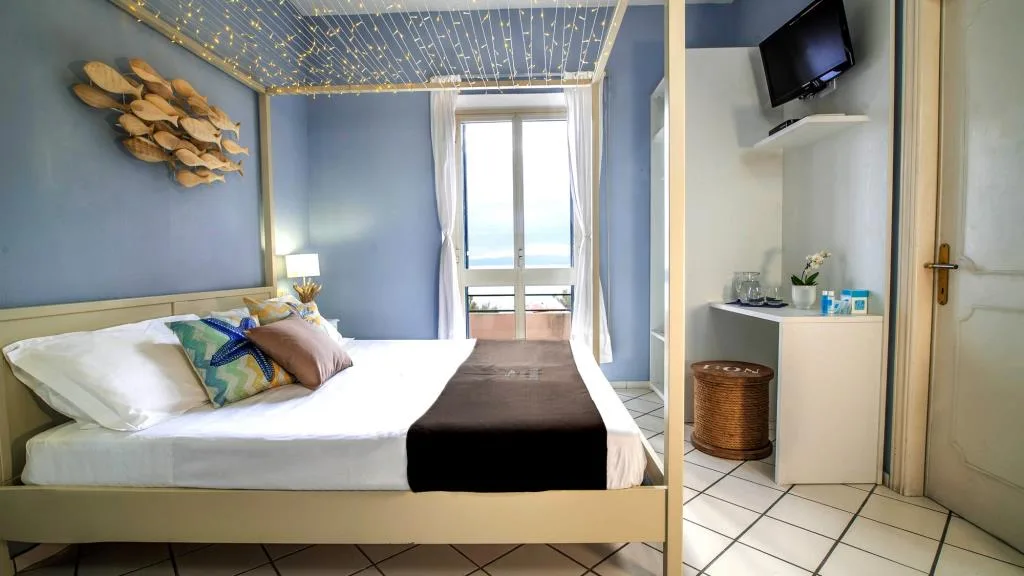 Camera azzurra con letto matrimoniale, luci a soffitto, TV e porta finestra con vista mare