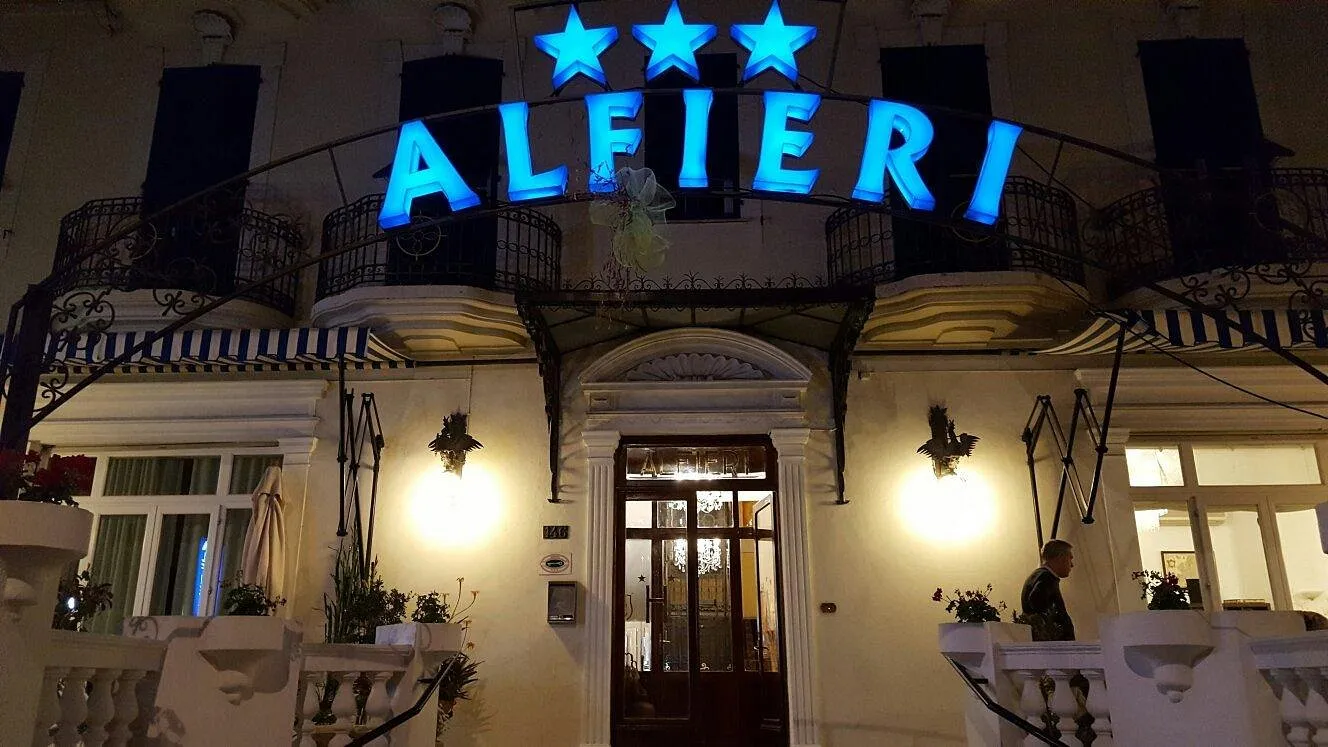 Ingresso notturno dell'hotel Alfieri con insegna luminosa blu e porta a vetri illuminata