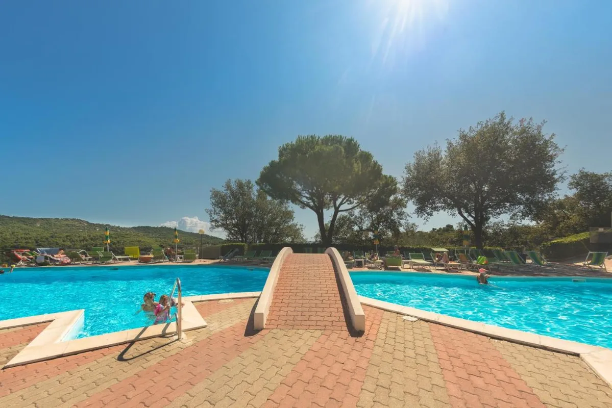 Piscina con ponticello e lettini circondata da alberi sotto il sole