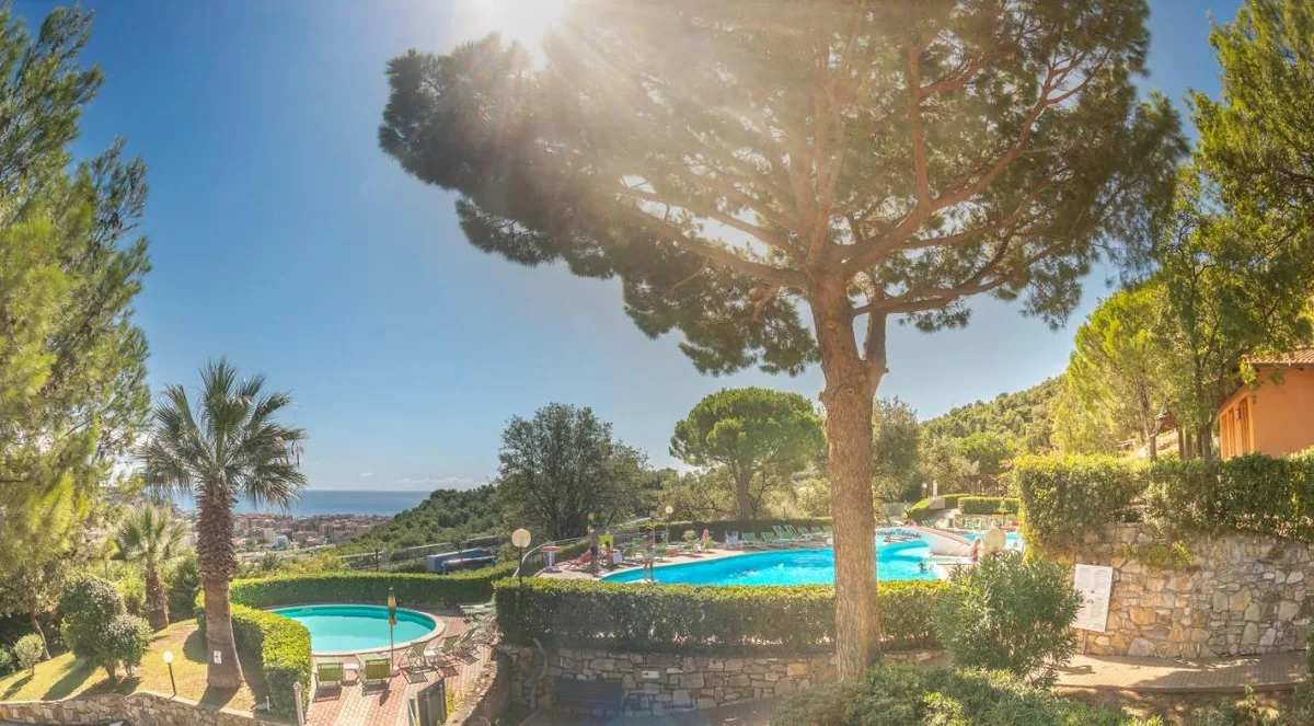 Vista panoramica sulle piscine del villaggio con pini, palme e mare sullo sfondo