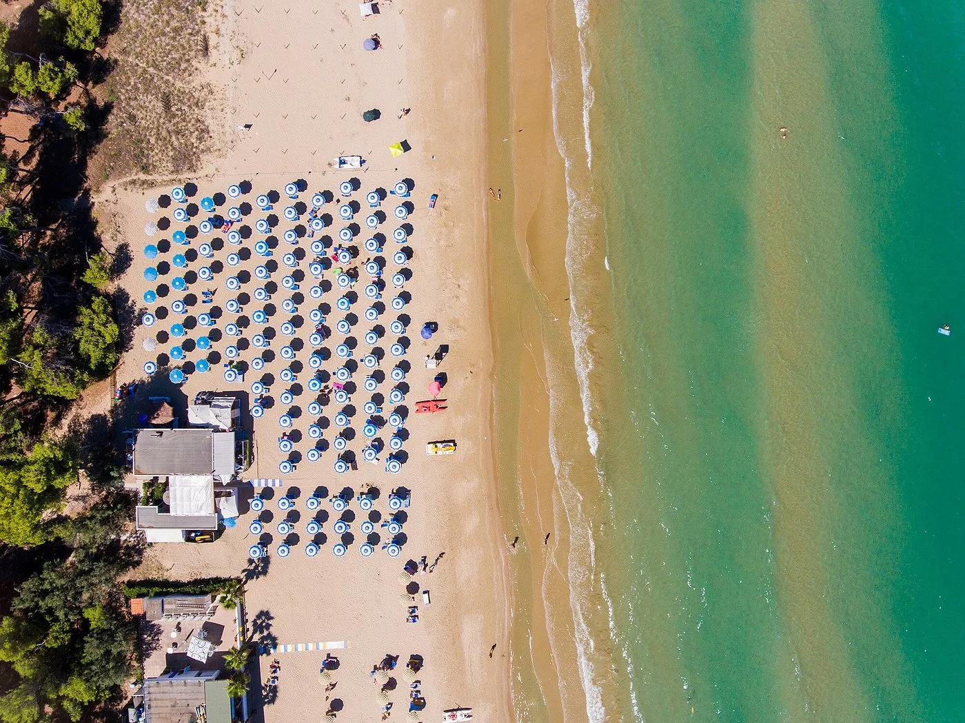 Vista aerea della spiaggia con ombrelloni blu allineati e mare turchese che lambisce la riva