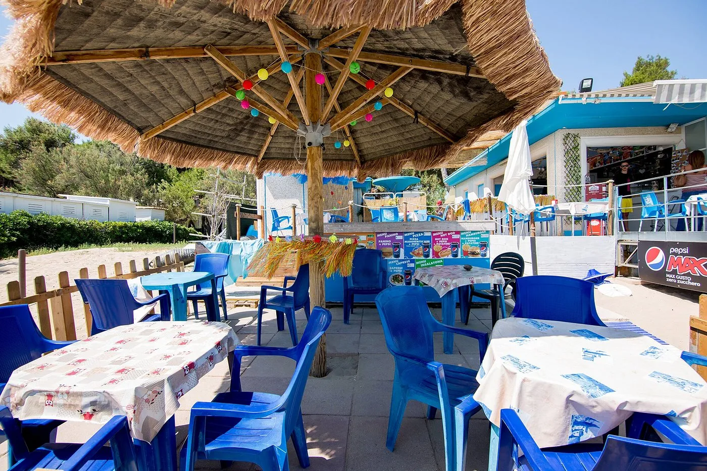 Area bar sulla spiaggia con ombrellone di paglia, tavoli e sedie blu e decorazioni colorate