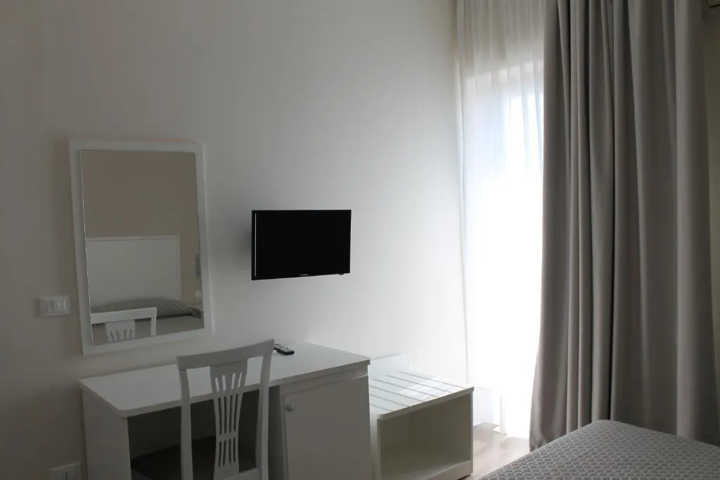 Camera con scrivania bianca, specchio, TV a parete e tende chiare