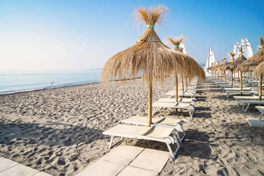 Spiaggia di sabbia con ombrelloni in paglia, lettini e barche a vela