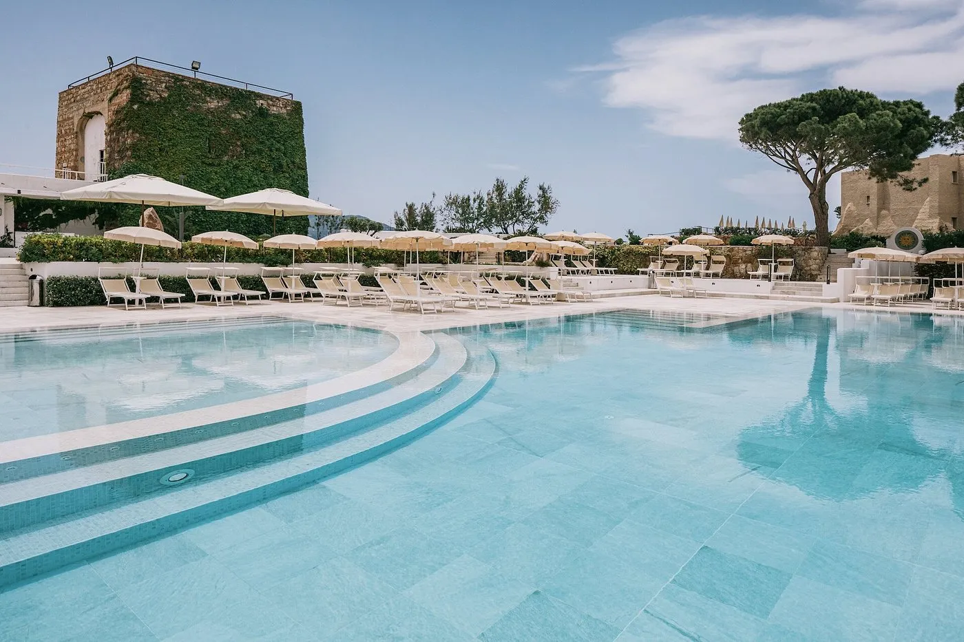 Piscina con acqua turchese, lettini e ombrelloni e torre in pietra sullo sfondo