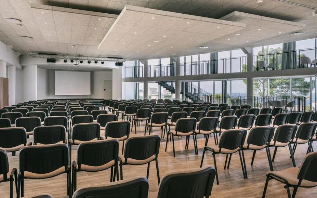 Sala congressi con file di sedie, schermo e ampie vetrate sul mare