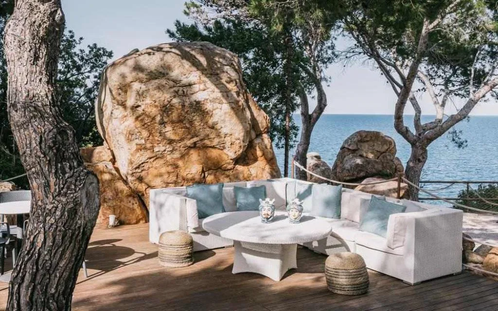 Salotto lounge esterno con divano bianco tra grandi rocce e pini vista mare