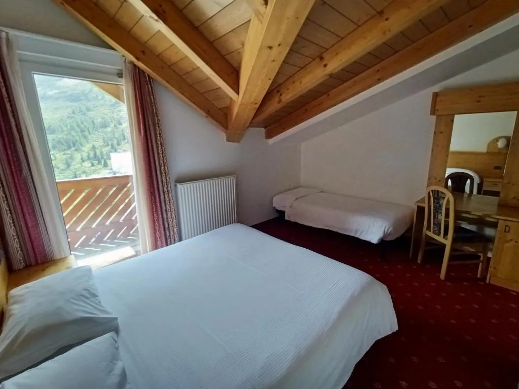 Camera mansardata con travi a vista in legno, letti e balcone con vista montagna
