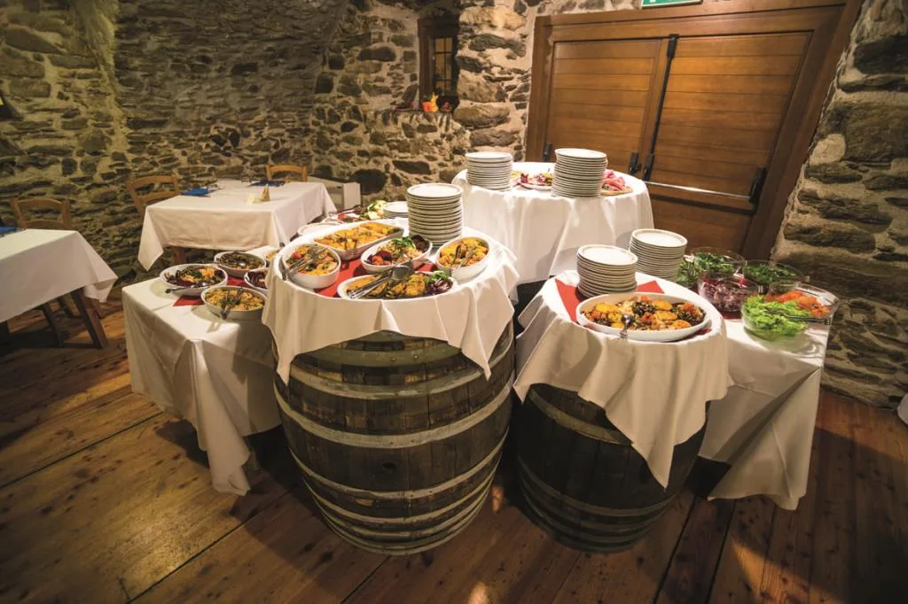 Buffet con piatti caldi e freddi disposti su botti in legno e tavoli con tovaglie bianche in sala dalle pareti in pietra