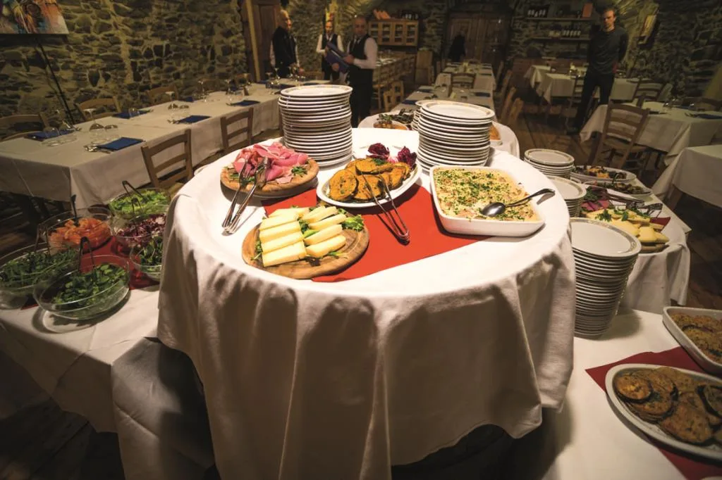 Tavolo buffet con salumi, formaggi e verdure, piatti impilati e personale di servizio sullo sfondo