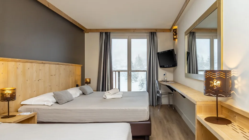 Camera dell'hotel con letto matrimoniale, testiera in legno, scrivania, TV e balcone con vista sulla neve