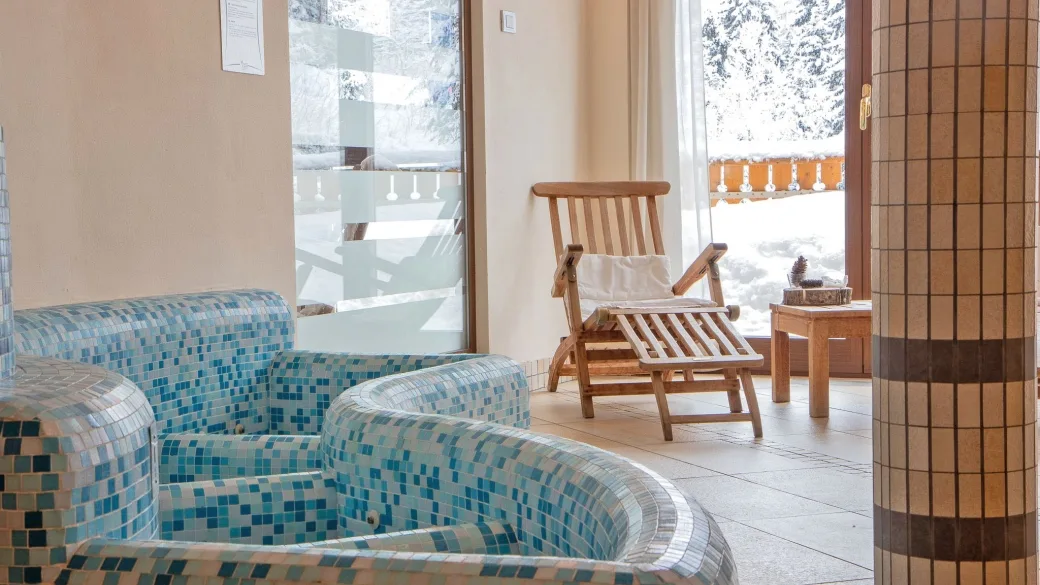 Area relax della spa con sedute in mosaico azzurro e lettino in legno vicino alle vetrate sulla neve